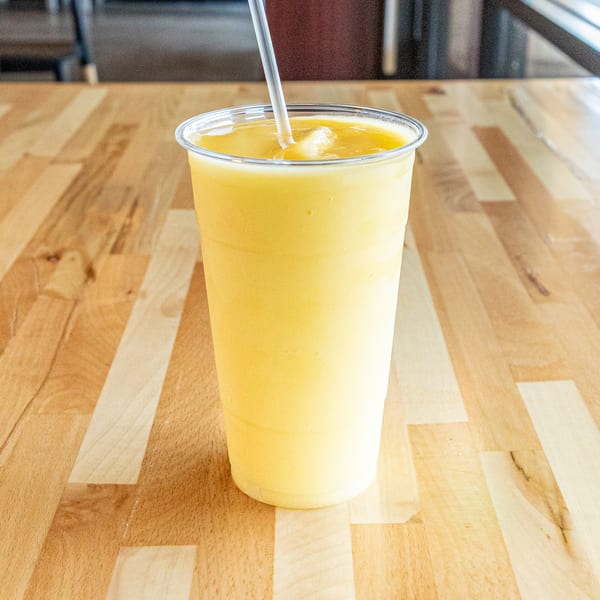 A tall plastic cup of yellow mango smoothie with a straw sits on a wooden table.