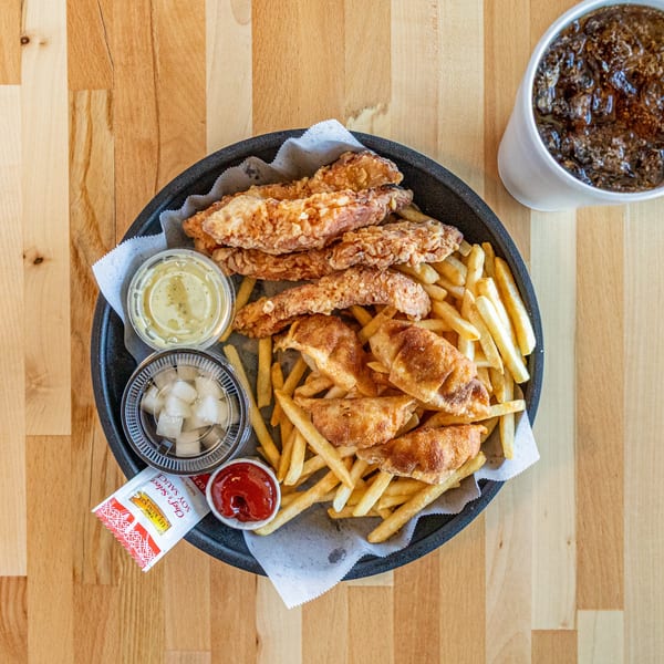 A black basket of fried chicken, french fries, dumplings, sauces, and a soda on a wooden table.