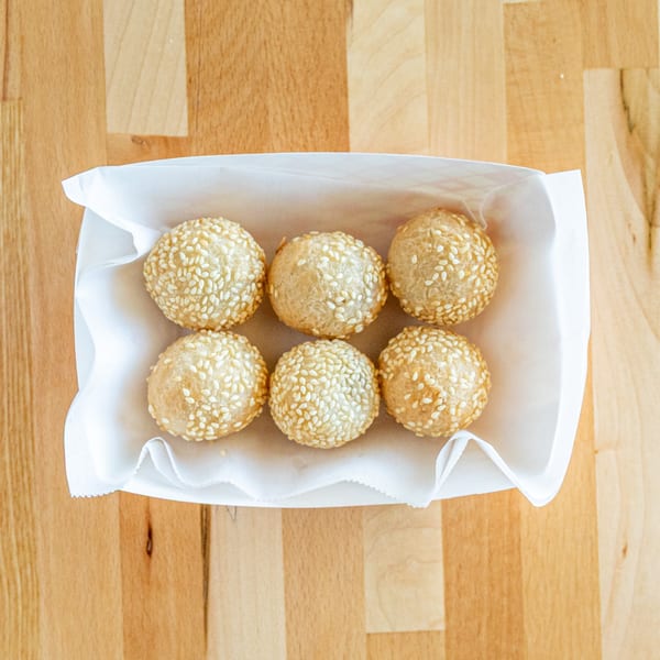 Six golden-brown sesame balls in a white paper tray on a light wooden table.