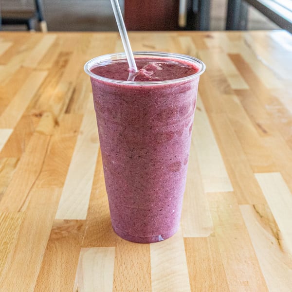 A vibrant purple berry smoothie with a straw in a clear plastic cup on a light wooden table.