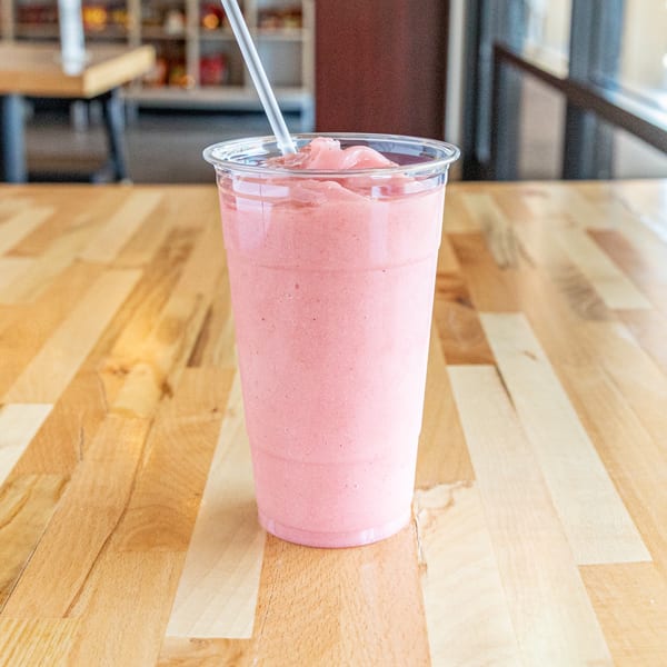 A pink strawberry milkshake in a clear plastic cup with a straw on a wooden table.